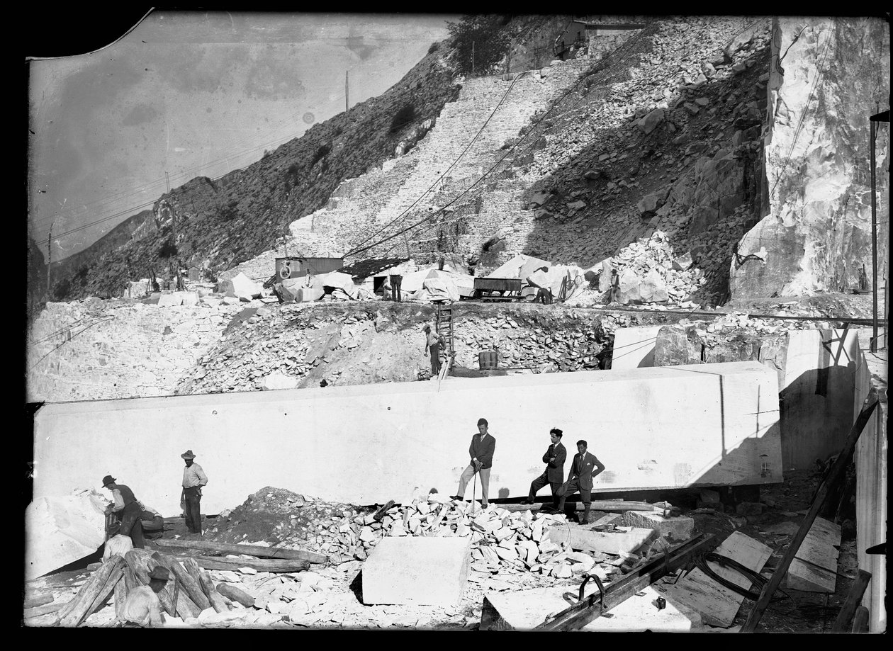 Drie mannen observeren het werk van verschillende graafmachines in de Apuane Alpen. Achter hen een enorm marmerblok, klaar om naar de vallei te worden vervoerd door A. Corsini