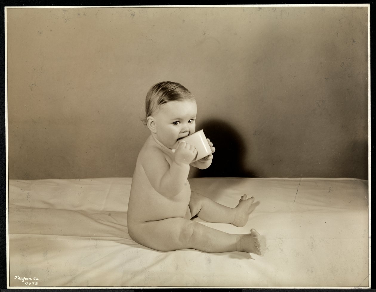 Een baby die in bed zit en drinkt uit een kopje bij het Maternity Center Association aan 70th Street East River, New York, 1935 door Byron Company