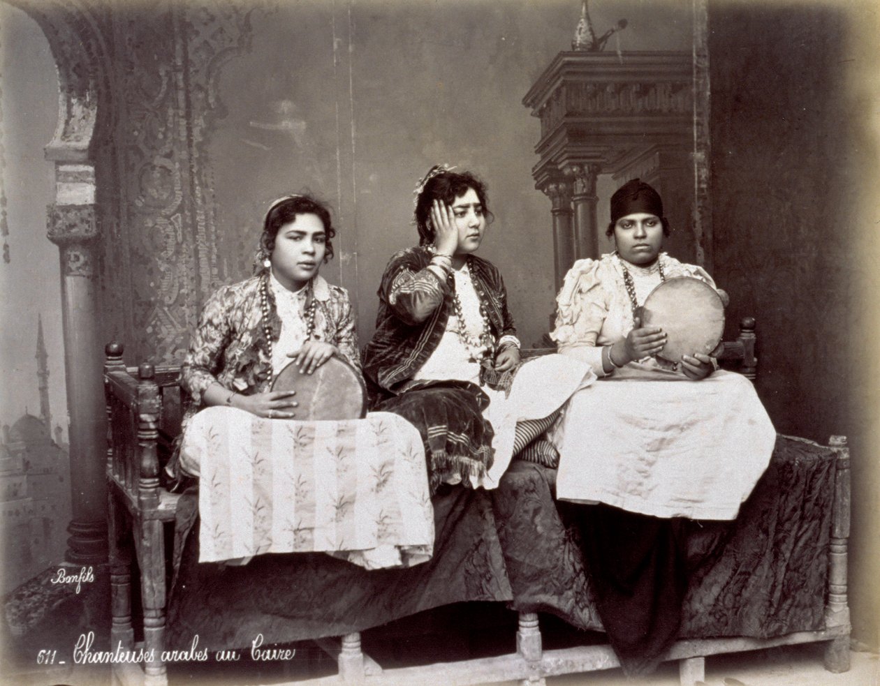 Portret van drie jonge Arabische zangers in etnische kleding met lange kettingen. Ze poseren in de studio, zittend. Twee van hen houden een tamboerijn vast door Félix Bonfils