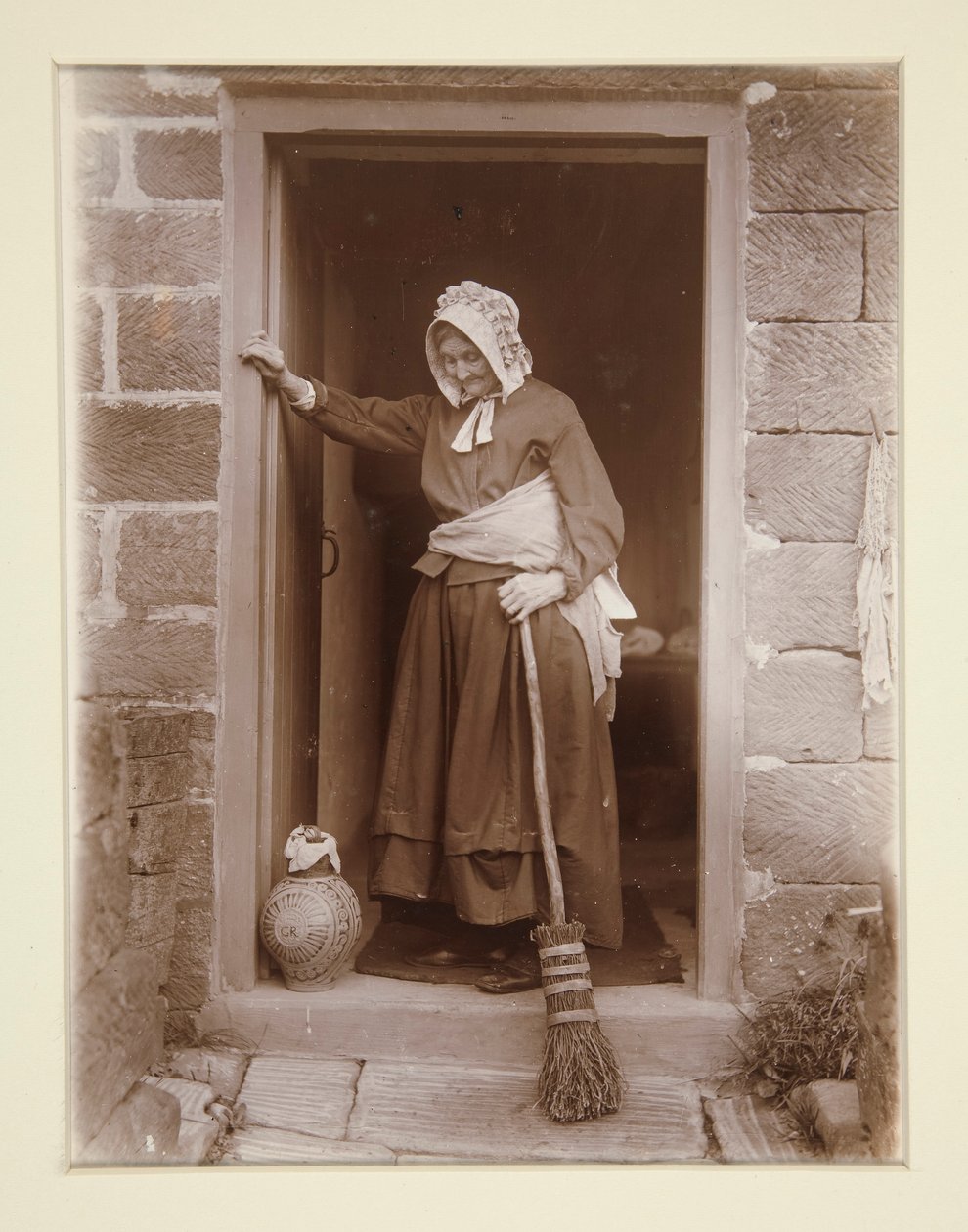 Een oude dame met een gistfles bij een cottage deur door Frank Meadow Sutcliffe