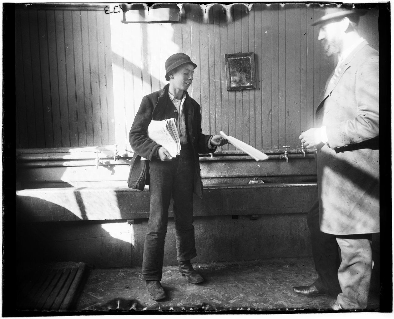 Portret van een krantenjongen die een krant overhandigt aan een man in wat lijkt op een toilet in het Duane Street Lodging House door Jacob August Riis