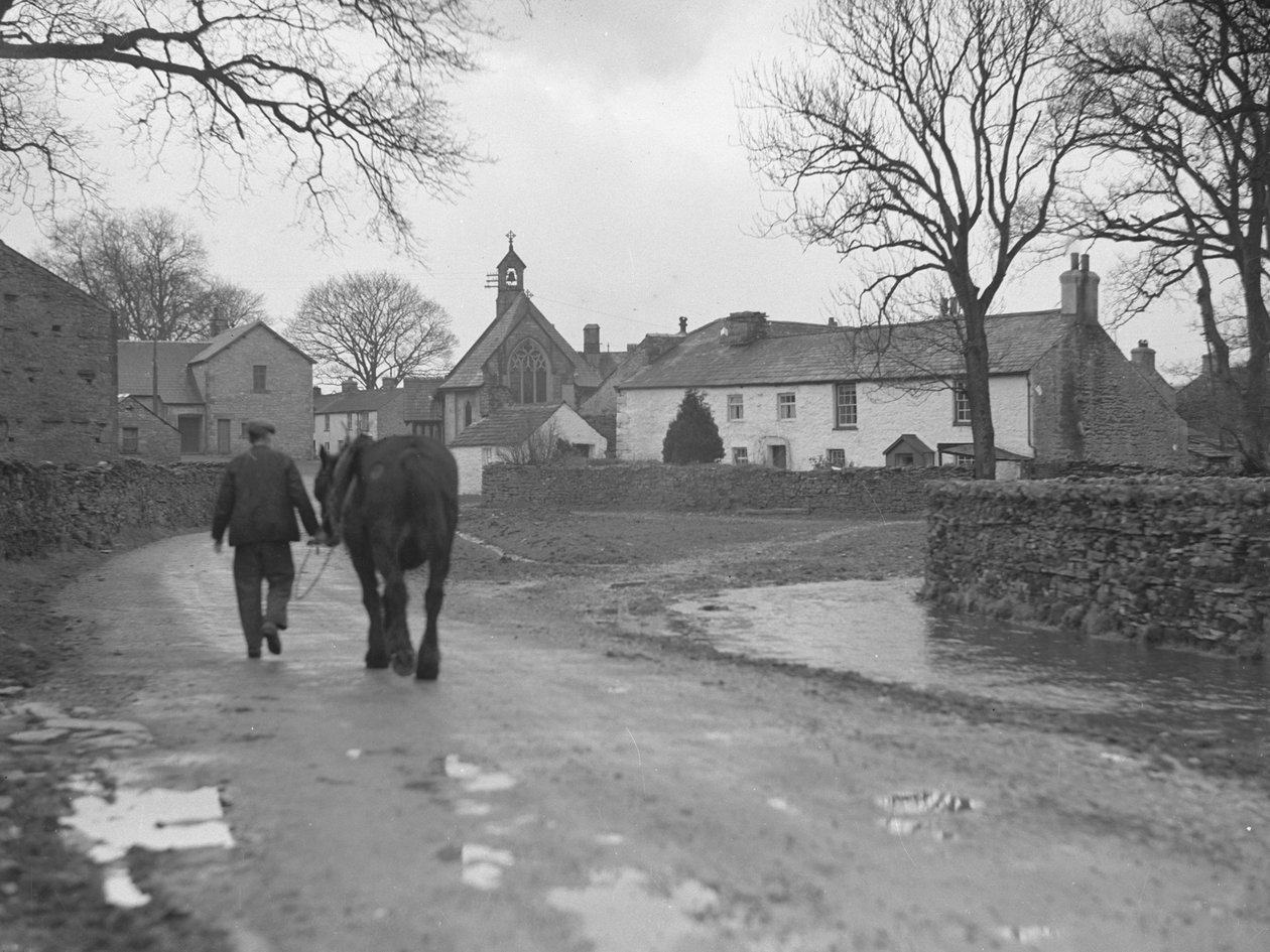 Man leidt paard over weg weg van camera naar dorpsgebouwen en kerk; architectuur; religieus; kerkelijk; door Joseph Hardman