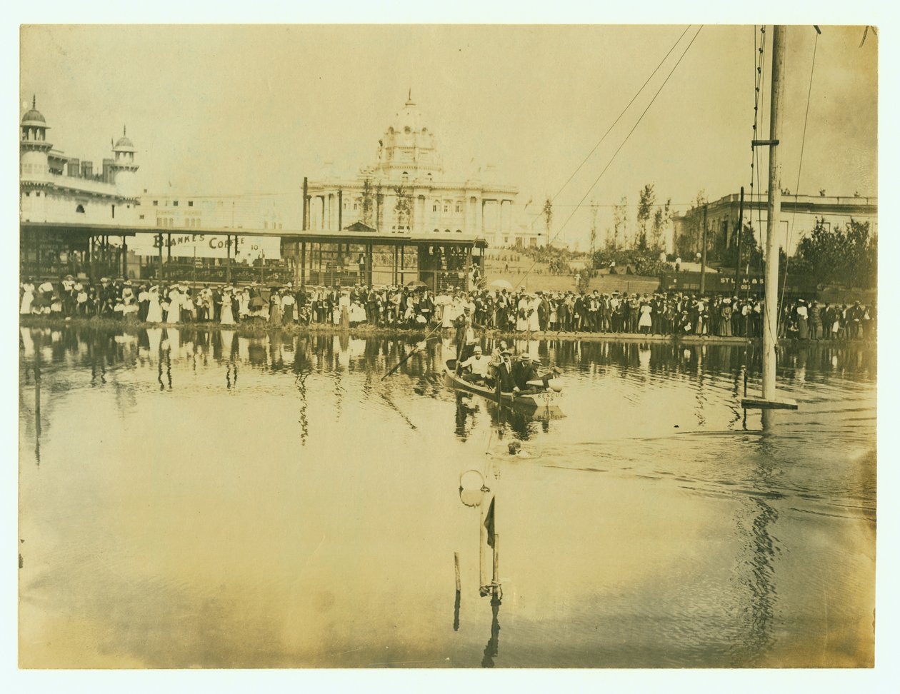 Horizontale foto van een zwemmer die een finishlijn passeert. Mannen in een boot kijken toe evenals menigten mensen aan de oever door Unknown