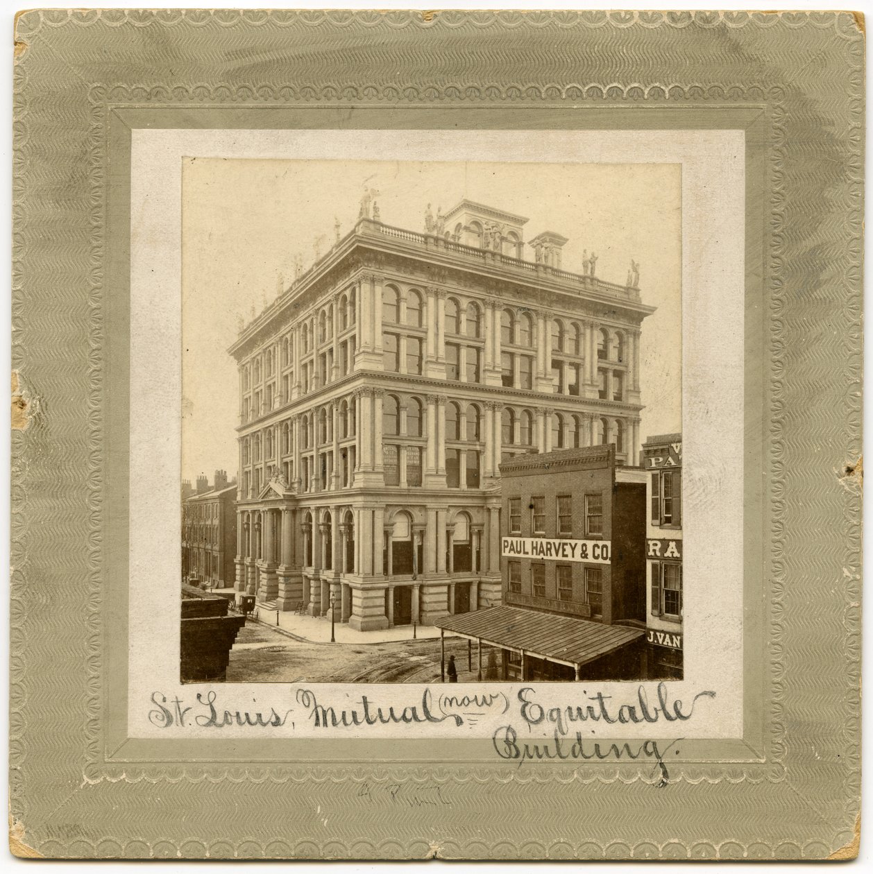Foto van een zes verdiepingen tellend gebouw met figuren langs de bovenkant. Er zijn andere gebouwen zichtbaar aan beide zijden. Gebouw ook wel St. Louis Life Insurance Building genoemd door Unknown