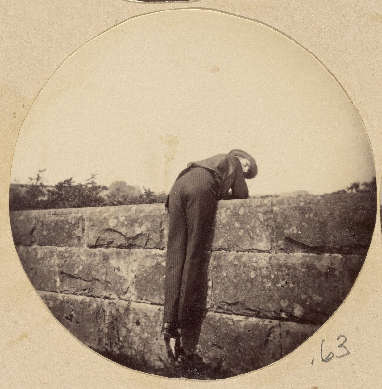 [Man met hoed gebogen over een stenen muur]. door Unknown artist