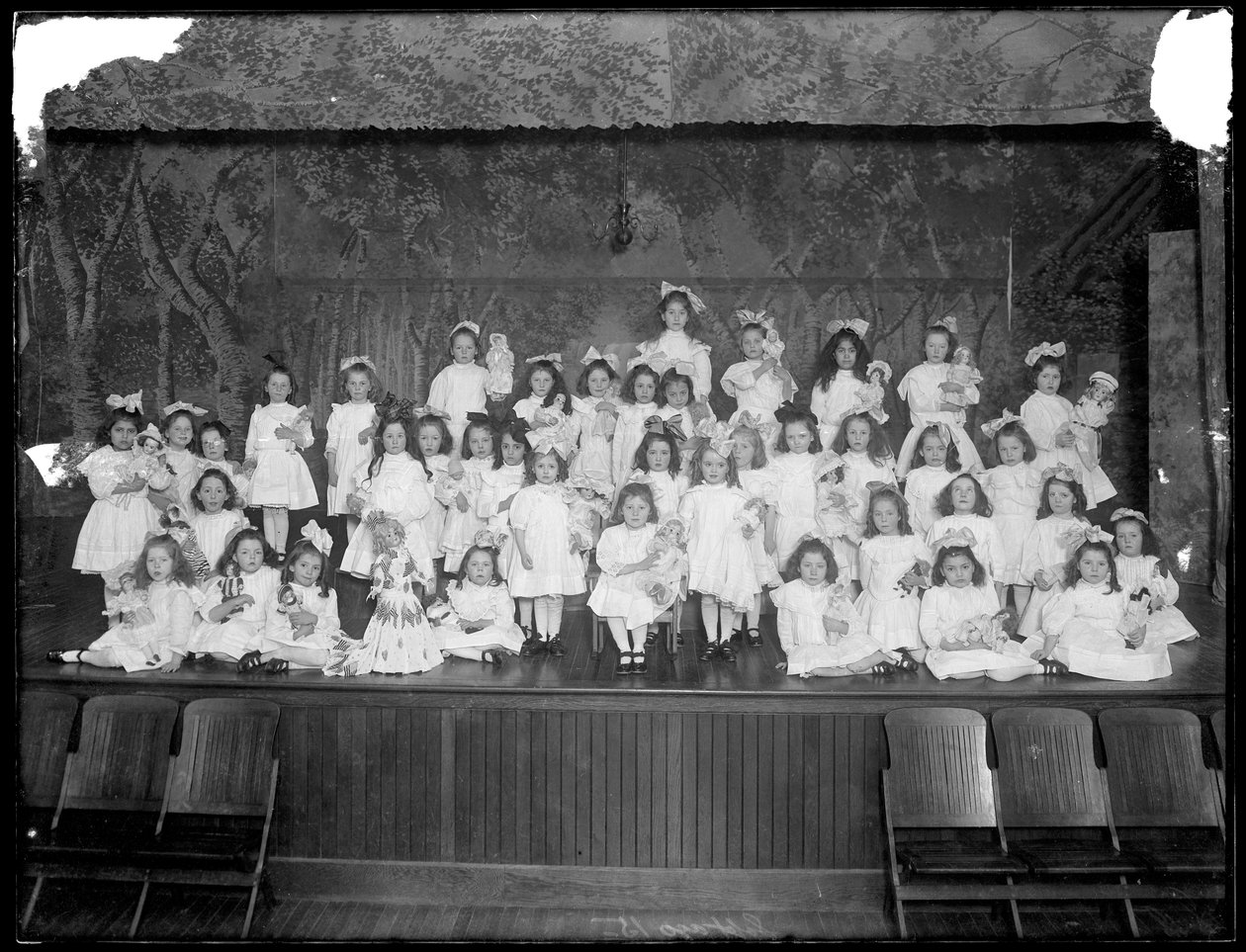 Groep kleine meisjes geposeerd op een klein podium met poppen, waarschijnlijk het Rooms-Katholieke Weeshuis, Kingsbridge, Bronx, ca. 1911 door William Davis Hassler