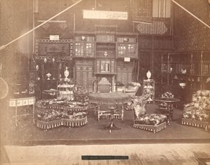 Tentoonstelling van S. Howard & Sons -- Hoofdgebouw, Collectie meubels, waaronder gestoffeerde stoelen, tafels, een open haard, kasten en schappen. Aan de muren hangen tapijten, en op tafels en planken staan vazen