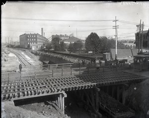Horizontale zwart-witfoto van een houten brug en tramsporen over gedeeltelijk aangelegde spoorlijnen. Een tram gemarkeerd Chouteau 2137 begint de brug over te steken aan de rechterkant. In de verte fabrieksgebouwen met de tekst Star door Charles Clement Holt