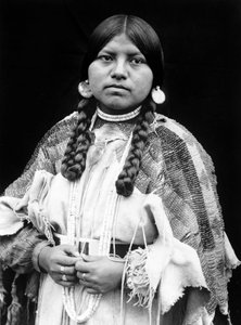 Cayuse vrouw, halflang portret, staand, naar voren gericht, vlechten, schelpschijf oorbellen, schelpkralen choker en schelpkralen om de nek, geborduurde hertenleren jurk met geborduurde riem c. 1910, foto Edward S. Curtis