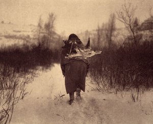 Op weg naar het kamp. Een apsaroke Indiaan die hout draagt in de sneeuw, in een klein winterkamp nabij een beek in de Pryor Mountains, Montana. Foto uit volume 4 van de encyclopedie gepubliceerd door Edward S. Curtis over de Indianen van Noord-Amerika.