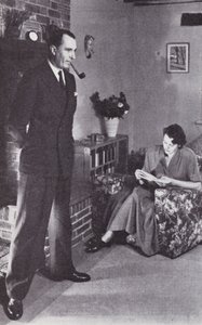 Vrouw leest, terwijl man zijn rug tegen het vuur verwarmt, ca. 1950 door English Photographer