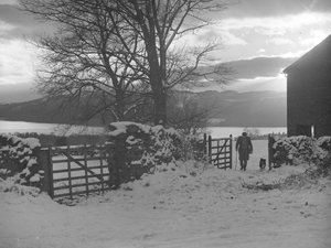 Man en hond lopen door hek naar camera in sneeuw met Thirlmere erachter door Joseph Hardman