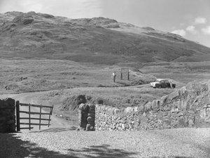 Zicht op de weg die over Hard Knott Pass gaat, twee mensen die bij een verkeersbord staan en een geparkeerde auto in de grasberm door Joseph Hardman
