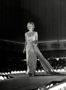 Model gefotografeerd op een catwalk in avondjurk met schouderbandjes van stras; het artikel is een creatie van het modehuis "Fumach".