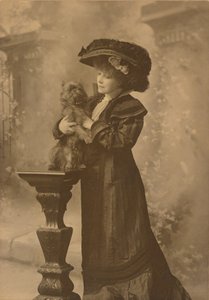 [Sarah Bernhardt staat in een studio en houdt een klein zwart hondje vast op een sierlijk, gebeeldhouwd houten voetstuk]. door Theodore C. Marceau