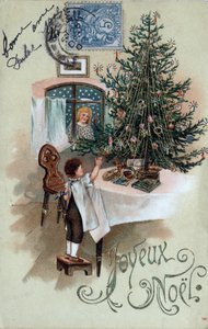 Kerstkaart: een kind voor een kerstboom poseert op een tafel, geobserveerd door een engel in het raam