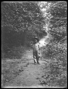 William Gray Hassler met Strohoed en Overall, met Hengel en Mand, wandelend op een Pad in het Bos