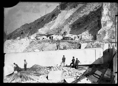 Drie mannen observeren het werk van verschillende graafmachines in de Apuane Alpen. Achter hen een enorm marmerblok, klaar om naar de vallei te worden vervoerd door A. Corsini