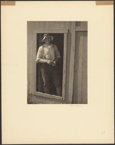[Man met strohoed staat in de deuropening van een schuur en houdt een tabaksbuidel vast]. door Doris Ulmann