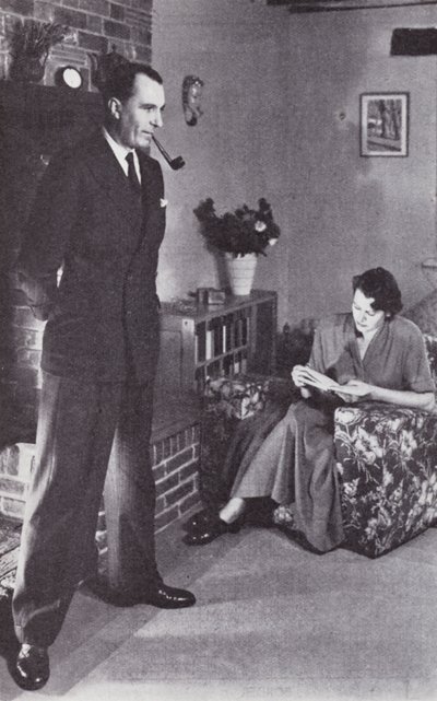 Vrouw leest, terwijl man zijn rug tegen het vuur verwarmt, ca. 1950 door English Photographer