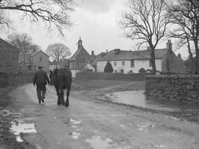 Man leidt paard over weg weg van camera naar dorpsgebouwen en kerk; architectuur; religieus; kerkelijk; door Joseph Hardman