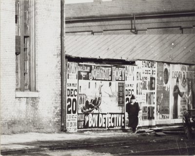 Een man loopt langs een muur met posteradvertenties, waaronder een voor Johnnie Hoey The Boy Detective in Havlin