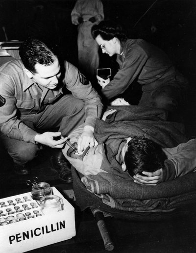 Tweede Wereldoorlog (1939-1945): Luxemburg februari 1945: In een evacuatieziekenhuis van het Amerikaanse leger dient een medicus (verpleegster) penicilline toe aan een gewonde soldaat om infectie te voorkomen terwijl een verpleegster hem voorbereidt op een door Unbekannt