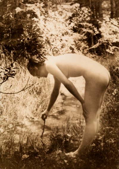 Een jonge vrouw, naakt in het bos, gezien in profiel terwijl ze zich bukt om een tak in een stroom te leggen door Unknown photographer