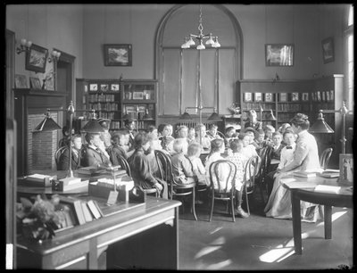 Kinderen luisteren naar een vrouw in de leeszaal van een niet-geïdentificeerde vestiging van de Queens Borough Openbare Bibliotheek, ca. 1910 door William Davis Hassler