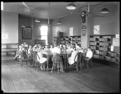Kinderen luisteren naar een vrouw in de leeszaal van een niet-geïdentificeerde vestiging van de Queens Borough Public Library, ca. 1910 door William Davis Hassler