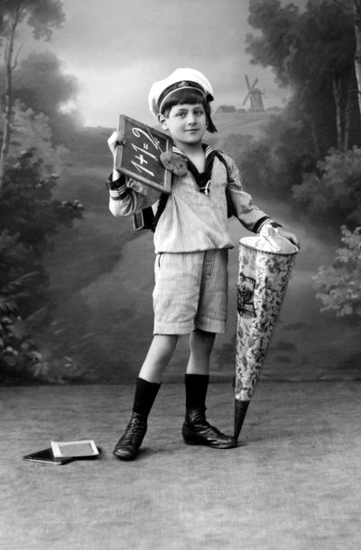 Eerste schooldag. Een jonge jongen in uniform poseert met een lei waarop de optelling 1+1=2 en een verrassingszakje. Fotografie rond 1910. door Ferdinand Georg Waldmuller