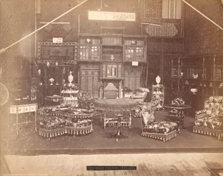 Tentoonstelling van S. Howard & Sons -- Hoofdgebouw, Collectie meubels, waaronder gestoffeerde stoelen, tafels, een open haard, kasten en schappen. Aan de muren hangen tapijten, en op tafels en planken staan vazen