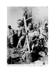Annie Jumper Tommie gebruikt een vijzel en stamper om maïsmeel te maken, en haar zoon, Tony Tommie, draagt een adelaarsveren tooi, 5 februari, 1927