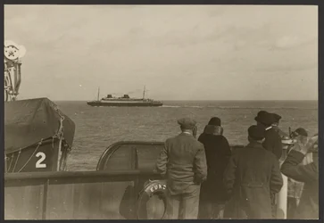 [Groep mensen op het dek van een schip die kijken naar een ander schip dat in de verte voorbij vaart].