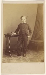 [Ongeïdentificeerd jongetje in pak staat bij een tafel met boeken erop].