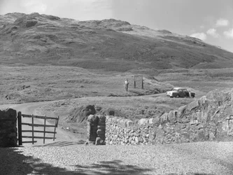 Zicht op de weg die over Hard Knott Pass gaat, twee mensen die bij een verkeersbord staan en een geparkeerde auto in de grasberm