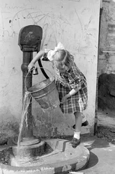 Klein meisje leegt een emmer water in een pompfontein, 9 mei 1939 (z/w foto)
